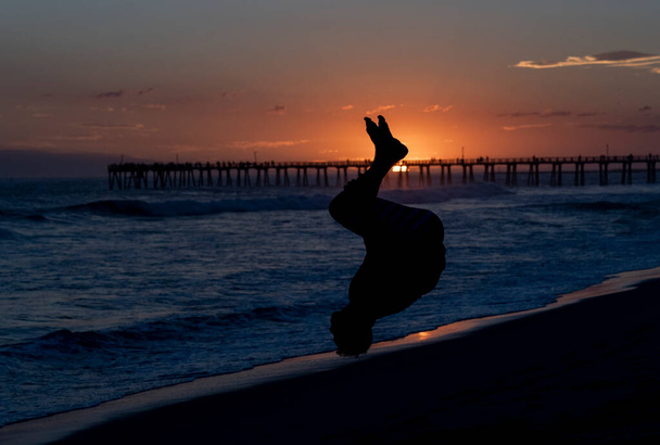 Назад фліп на пляжі на заході сонця з пірсом на фоні
 - Фото, зображення