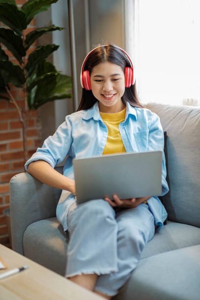 Mujer asiática joven estudiando, aprendiendo en línea, y escuchando música con auriculares mientras está sentada en el sofá en casa. Escuchar podcast, audiolibro, sonreír, y, viendo videos usando laptop. - Foto, imagen