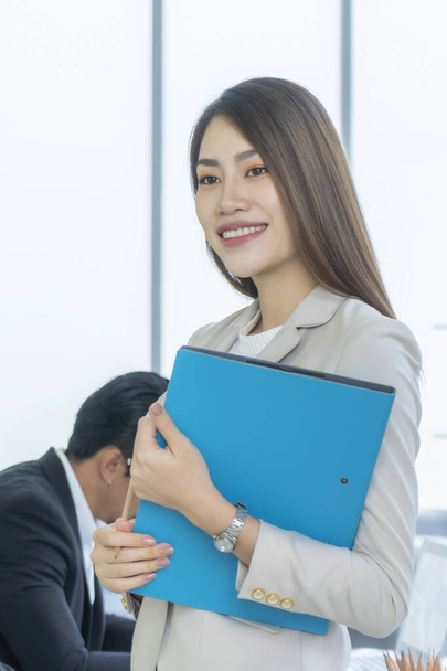 Mooie Aziatische zakenvrouw staat in de buurt van glazen raam met blauwe document map in haar hand. Tijdens haar werkuren. Ze glimlachte gelukkig.. - Foto, afbeelding