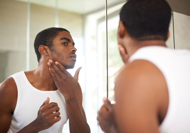 Er pflegt seine Haut. Ein junger Mann cremt sich sein Gesicht ein, während er in den Spiegel schaut - Foto, Bild