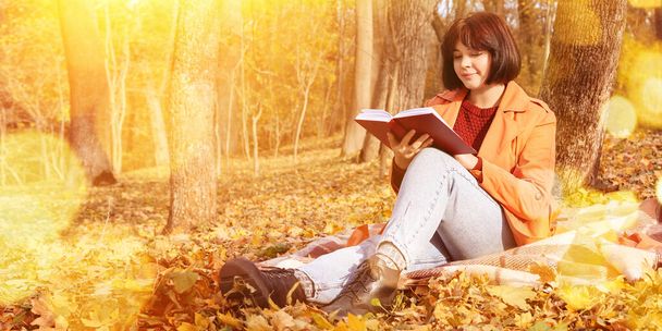 Genç bir kadın, sonbahar parkında ağaçların yanında oturmuş kitap okuyor. - Fotoğraf, Görsel