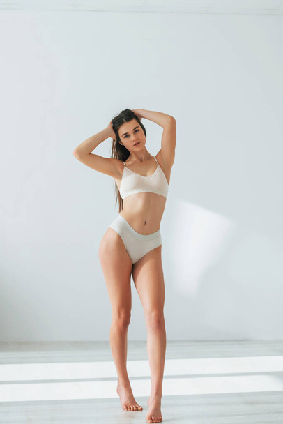 Standing in white clothes. Beautiful woman in underwear is posing indoors. - Foto, imagen