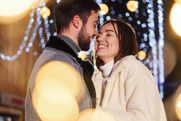 Beautiful garland lighting. Happy young couple celebrating New year outdoors on the street. - Φωτογραφία, εικόνα