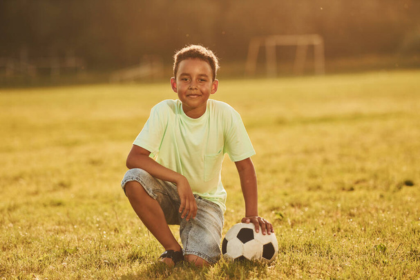 Sits with soccer ball. African american kid have fun in the field at summer daytime. - Zdjęcie, obraz