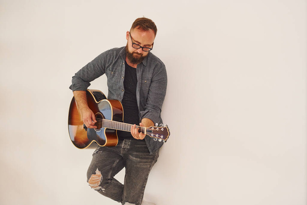 Standing in the studio. Man in casual clothes and with acoustic guitar is indoors. - Foto, Bild