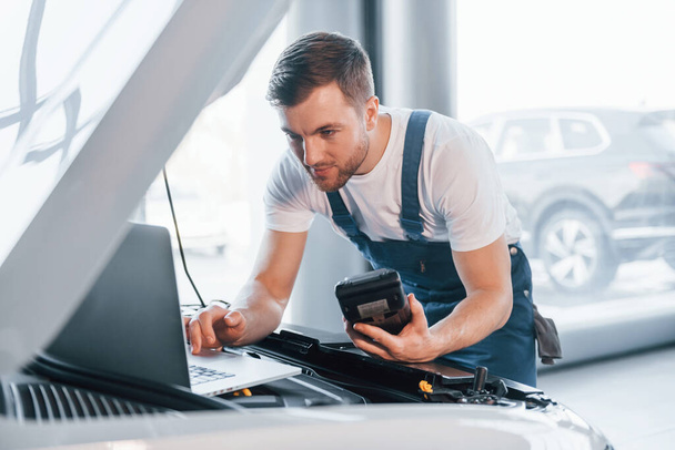 Uses laptop. Young man in white shirt and blue uniform repairs automobile. - Photo, Image