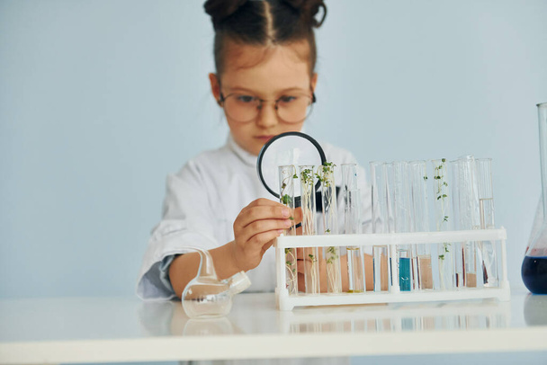 Holds magnifying glass. Little girl in coat playing a scientist in lab by using equipment. - Foto, afbeelding