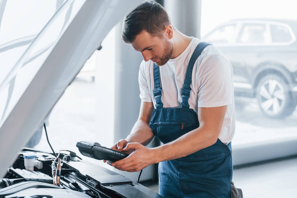 Young man in white shirt and blue uniform repairs automobile. - 写真・画像