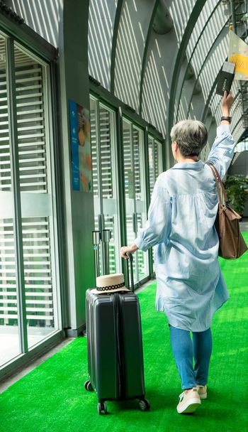 Retrato de adulto mayor anciano asiático mujer de 60 años sonriendo de pie con maleta maleta de equipaje y sosteniendo bandera guiada poste para el concepto de viaje. - Foto, imagen