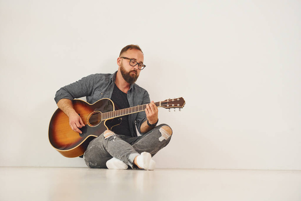 In the studio. Man in casual clothes and with acoustic guitar is indoors. - Foto, afbeelding