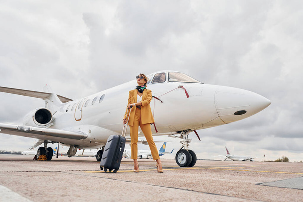 Elegant clothes. Young woman with luggage is outdoors near airplane. - Fotó, kép