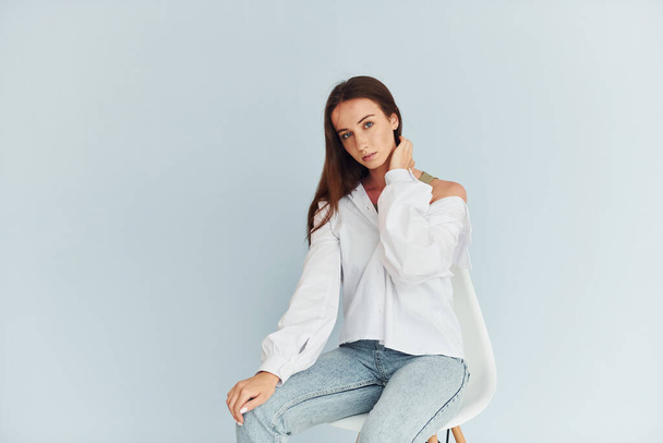Young woman in white clothes sitting indoors against white background. - Valokuva, kuva
