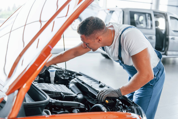 Orange colored car. Man in uniform is repairing broken automobile indoors. - Фото, изображение