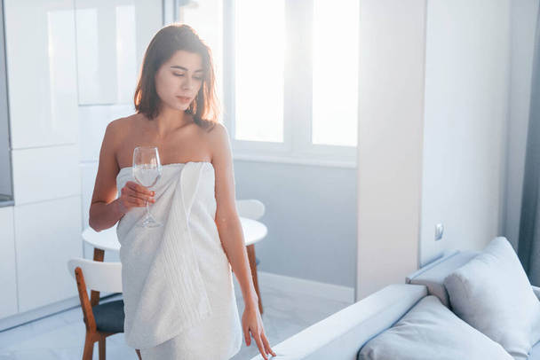 In white towel. Young woman is indoors in room of modern house at daytime. - Foto, Imagem
