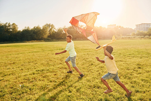 Running with kite. Two african american kids have fun in the field at summer daytime together. - 写真・画像