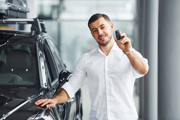 Holds keys. Young man in white shirt is indoors with modern new automobile. - Fotoğraf, Görsel