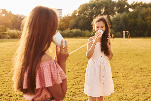 Kids having fun with tin can telephone. Standing outdoors on the sportive field. - 写真・画像