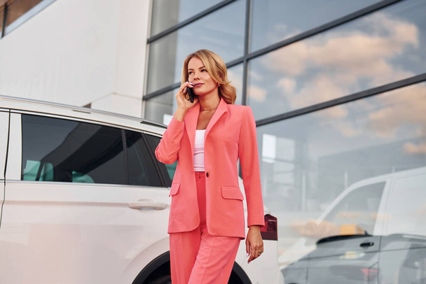 Having conversation by phone. Woman in formal wear standing outdoors with phone in hands and near car. - Photo, image