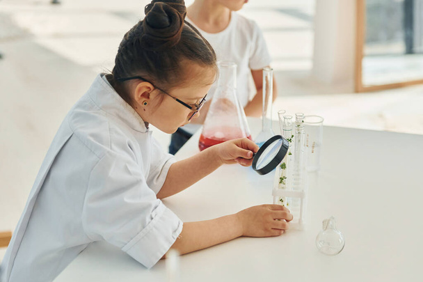 Holds magnifying glass. Little girl in coat playing a scientist in lab by using equipment. - Fotografie, Obrázek