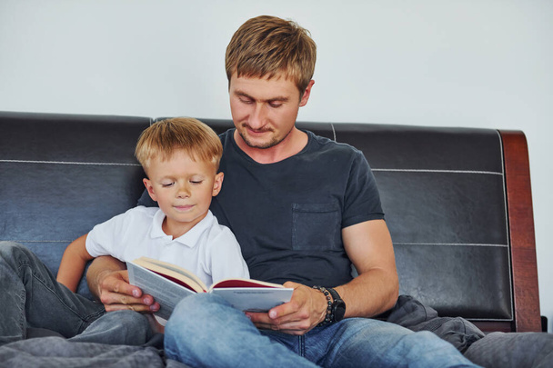With book in hands. Father and son is indoors at home together. - Photo, Image