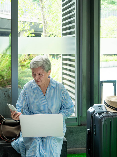 Расслабьтесь, отпуск, путешествия, портрет пожилой пожилой азиатской женщины 60-х годов улыбаясь с помощью компьютера ноутбука и смартфона в ожидании своего времени путешествия. - Фото, изображение