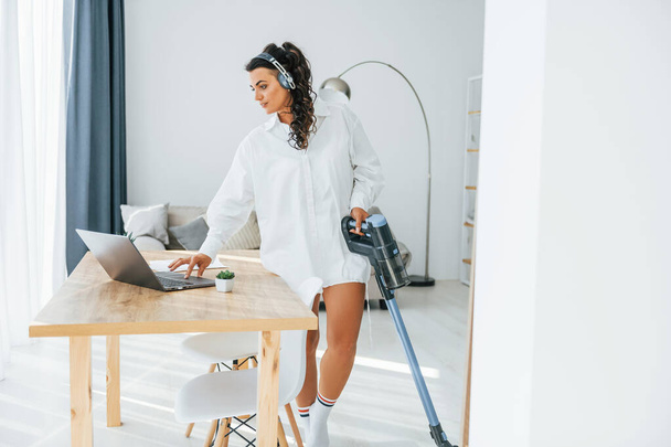 In headphones and with vacuum cleaner. Woman in casual clothes is indoors at home. - Foto, Imagen
