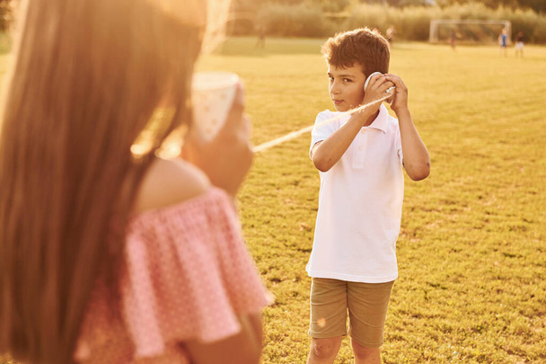 Kids having fun with tin can telephone. Standing outdoors on the sportive field. - Foto, Imagen