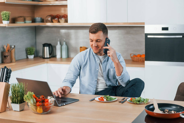 Talking by phone. Man preparing food at home on the modern kitchen. - Foto, afbeelding