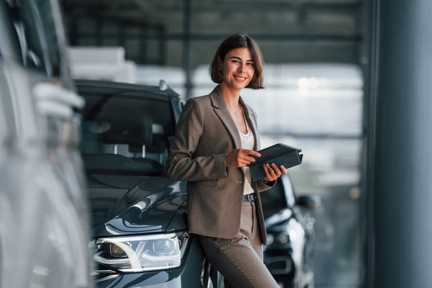 With tablet in hands. Woman is indoors near brand new automobile indoors. - Foto, Bild
