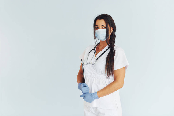 Standing against white background. Young female doctor in uniform is indoors. - Фото, изображение
