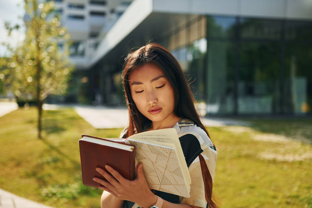With book in hands. Young asian woman is outdoors at daytime. - Photo, image