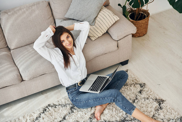 With laptop. Beautiful young woman in jeans and white shirt is at home. - Foto, Bild