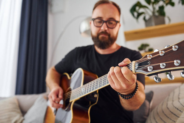 Playing the instrument. Man in casual clothes and with acoustic guitar is indoors. - Fotoğraf, Görsel