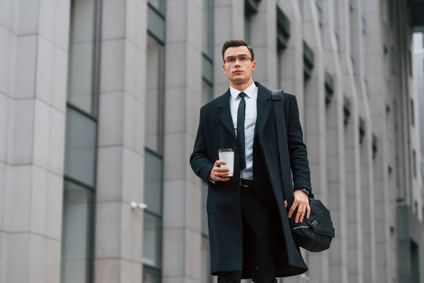 With cup of drink. Businessman in black suit and tie is outdoors in the city. - Foto, afbeelding