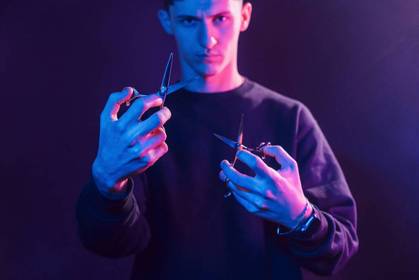 Holds scissors. Young barber with work equipment standing in the studio with neon lighting. - Фото, изображение