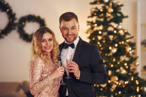 Standing against christmas tree. Happy couple have a new year party indoors together. - Photo, Image