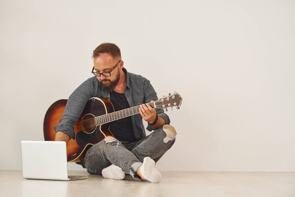 With laptop. Man in casual clothes and with acoustic guitar is indoors. - Foto, Bild