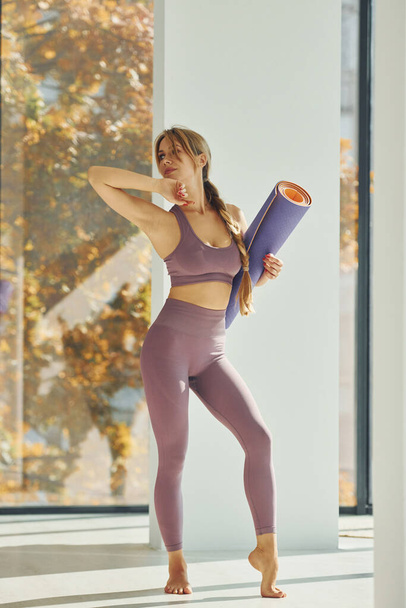 Standing and holding mat. Woman in sportive clothes doing yoga indoors. - 写真・画像