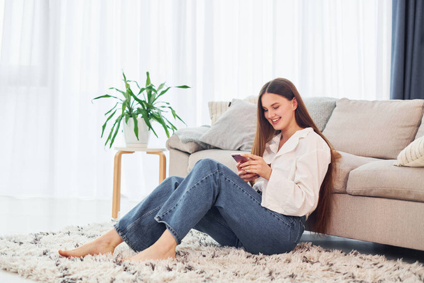 Sits near sofa with phone in hands. Young woman in white shirt and jeans is at home. - Foto, Bild