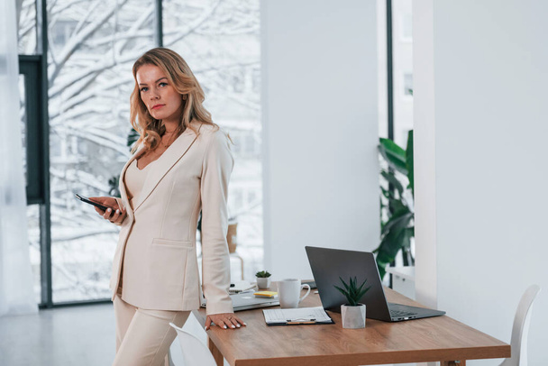 Standing near window. Business woman in formal clothes is indoors posing for the camera. - Foto, afbeelding