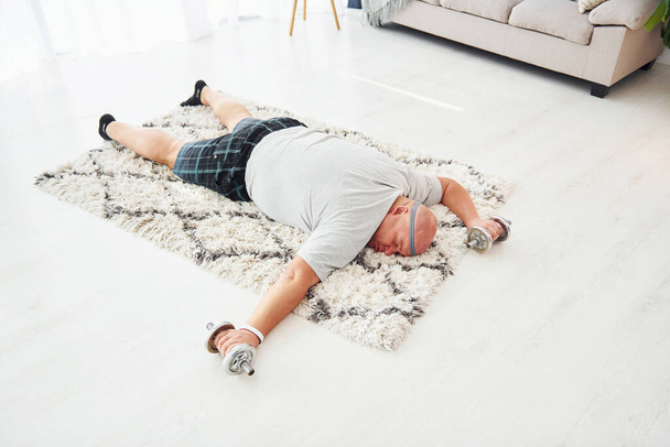 Laying down exhausted. With dumbbells in hands. Funny overweight man in casual clothes is indoors at home. - Fotoğraf, Görsel