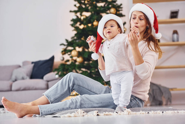 In Santa hats. Mother with her little daughter is indoors at home together. - Φωτογραφία, εικόνα