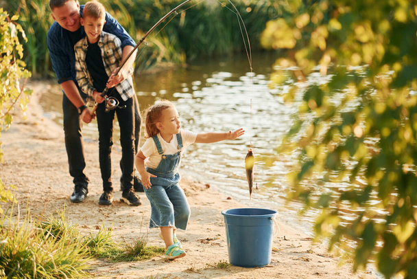 Girl putting fish into the bucket. Father with son and daughter on fishing together outdoors at summertime. - 写真・画像