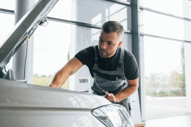 Repairing of the car. Man in uniform is working in the autosalon at daytime. - Zdjęcie, obraz