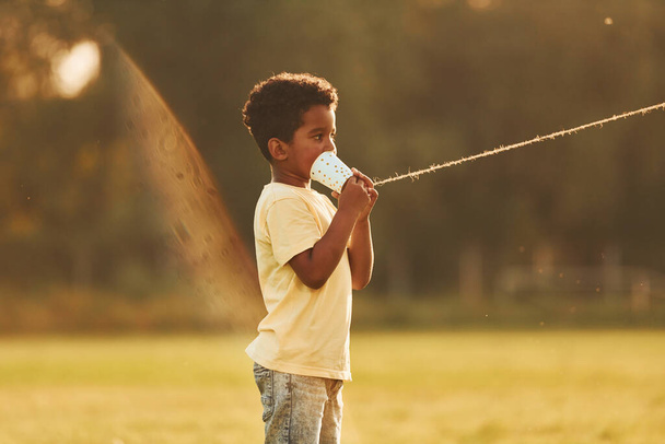 Talking by using cup on knot. African american kid have fun in the field at summer daytime. - Foto, immagini