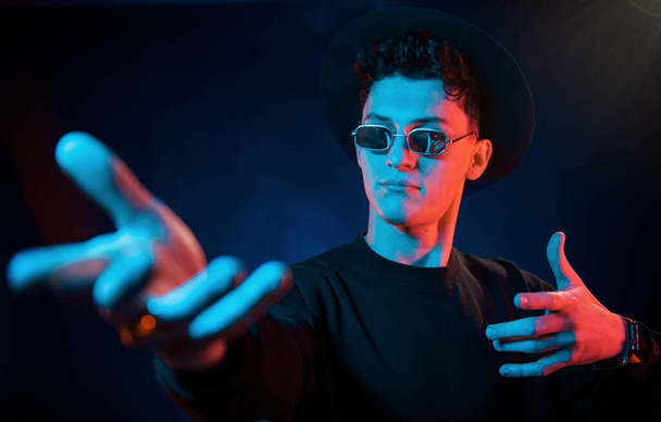 In hat and glasses. Neon lighting. Young european man is in the dark studio. - Foto, Imagem