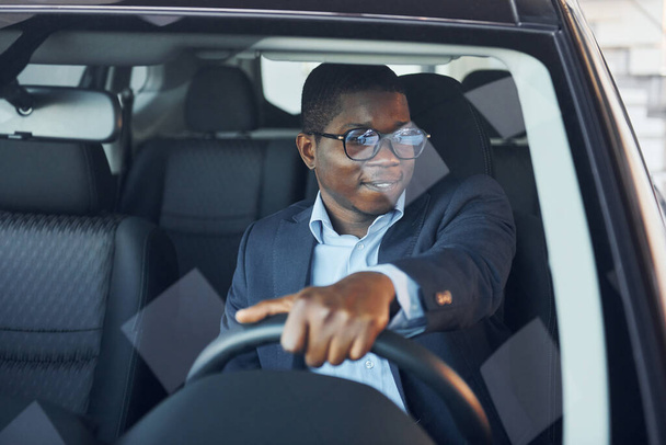 Front view. Young african american businessman in black suit is in the automobile. - 写真・画像