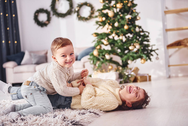Makaamassa ja pelaamassa. Pikkuveli ja sisko ovat joulun koristellussa huoneessa yhdessä. - Valokuva, kuva
