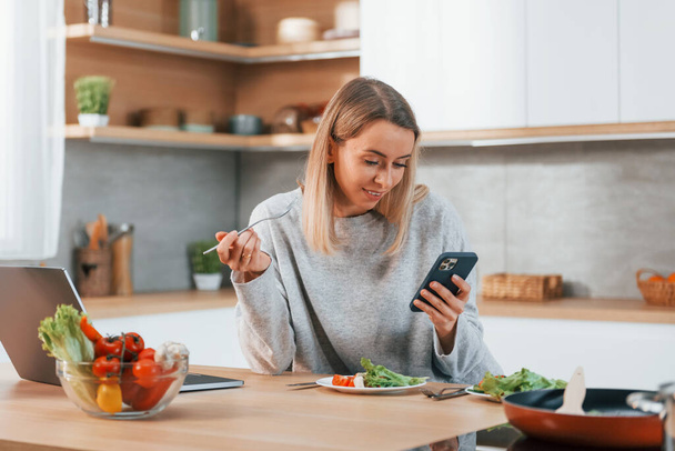Holding phone. Woman preparing food at home on the modern kitchen. - Photo, image