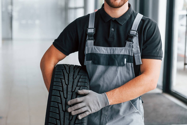 Holding tire. Man in uniform is working in the autosalon at daytime. - Φωτογραφία, εικόνα